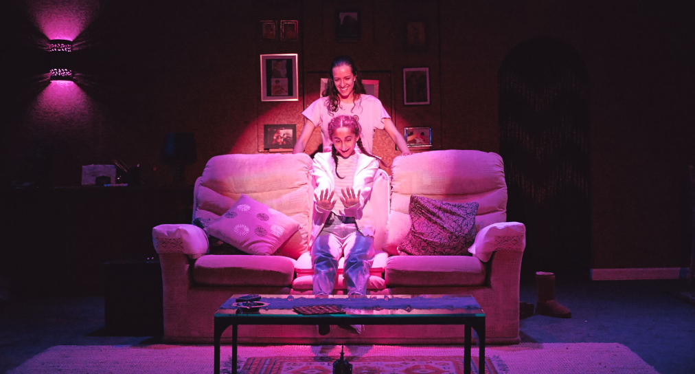 a photo from the play Favour, a young girl is sitting on a sofa admiring her manicure, her mother stands behind her smiling. The lighting is pink.