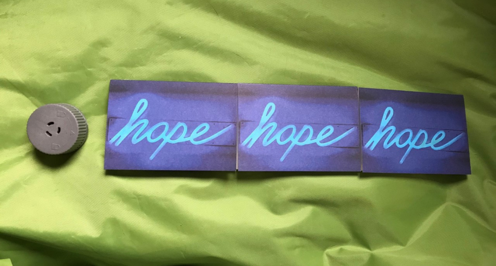 3 postcards which say the word 'hope' in neon lights, laid out of a green sheet