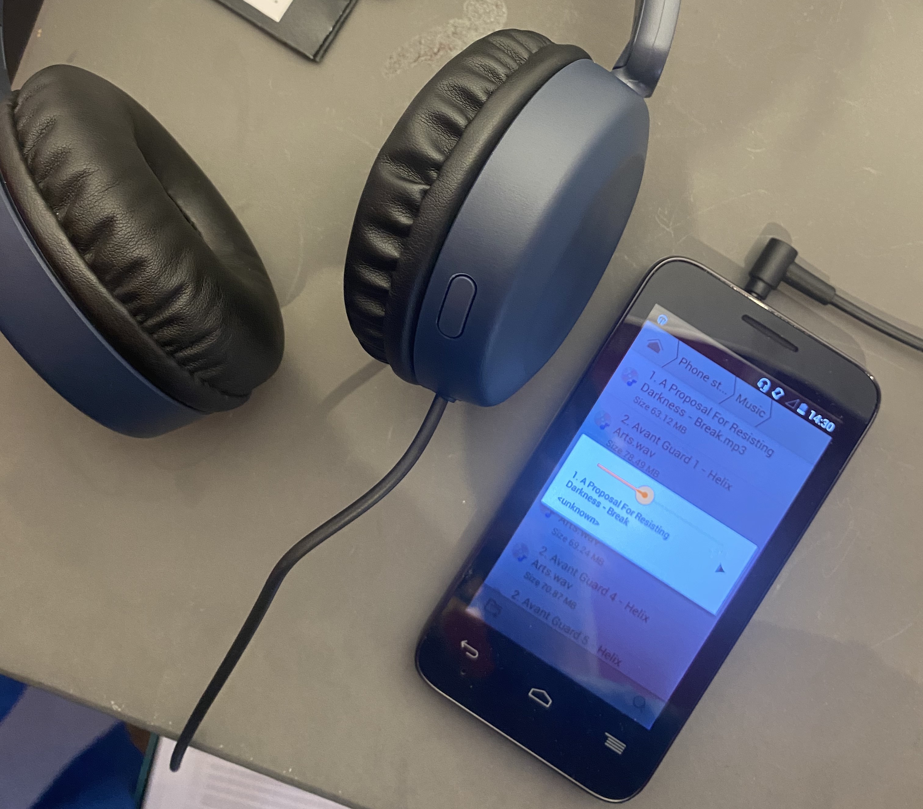 An MP3 player is shown on a grey surface, alongside grey headphones, with an audio file playing titled 'A Proposal for Resisting Darkness'