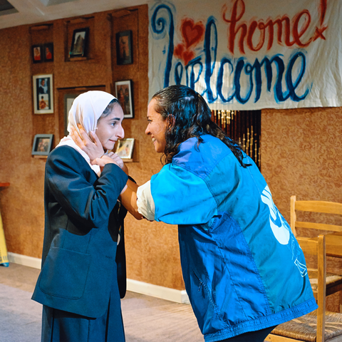 a production photo for Favour, Aleena is holding Leila's face in her hands and looking at her. In the background a banner reads 'Welcome home
