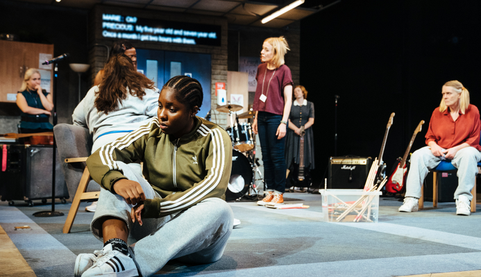 The Typical Girls cast on stage, Lara Grace Ilori is closest to the camera, she is sat on the floor with her legs crossed, looking at the floor.