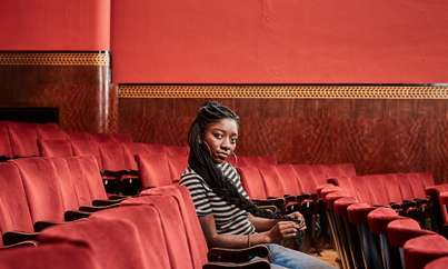 A photo of Eno Mfon sitting in a theatre and looking at the camera. Her hair is in long braids.