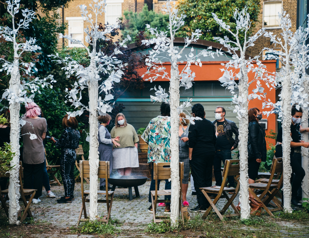 a photo from our production of Through This Mist. In the Clean Break garden, audience members are standing around and talking, wearing masks, standing around a circle of white make shift trees