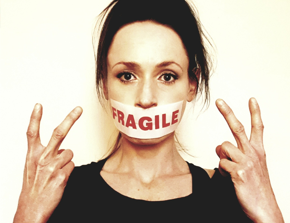 a photo of Sarah Cowan looking straight at the camera with 'fragile' tape over hear mouth, she is holding 2 fingers up with both hands.