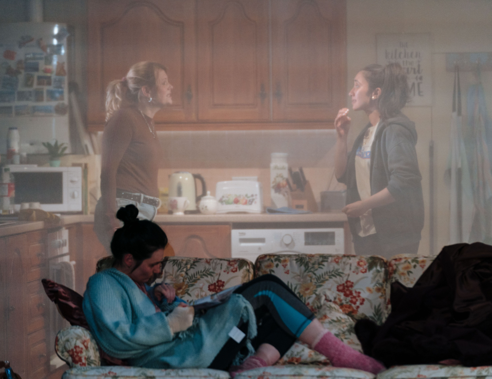 a photo from the set of Dixon and Daughters. A woman is lounging on a sofa, through a translucent wall you can see into a kitchen where two women are having a heated discussion.