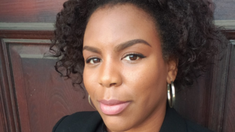 a headshot of Nola Sterling, standing in front of a door wearing a black blazer