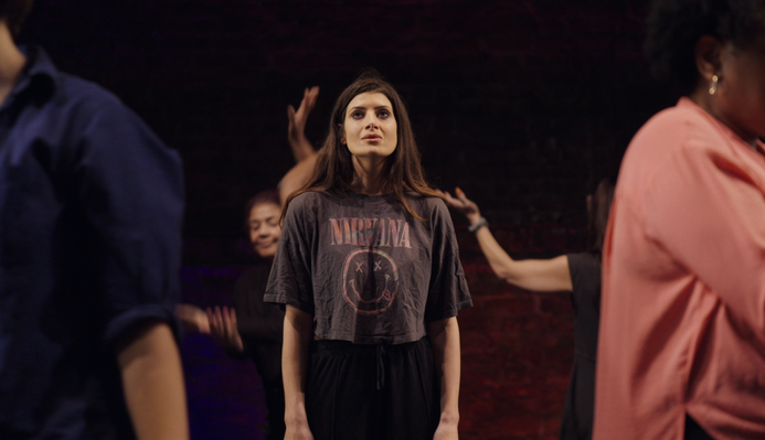 Natasha on a stage, looking upwards, surrounded by Clean Break Members
