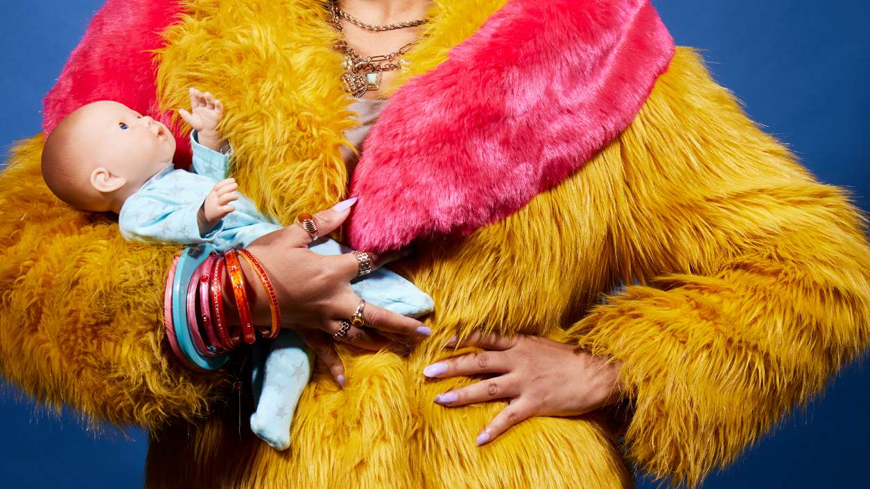 A woman's torso, wearing a brightly coloured fur coat, holding and baby doll