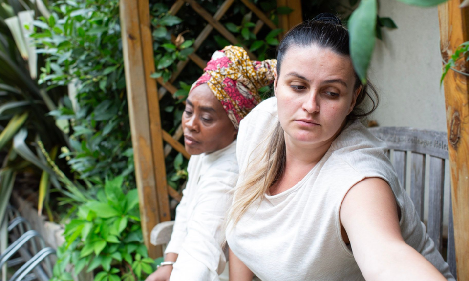 Two women sit in the Clean Break courtyard on a bench, performing as part of a dance sharing. They are stretching their arms out to their left and looking down.