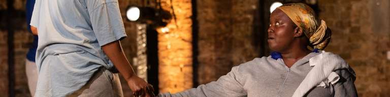 Two women stand on stage. They both wear loose grey clothes. The woman on the left has her back to the viewer, and is stretching her hand towards the other woman, who is seated and facing towards us.