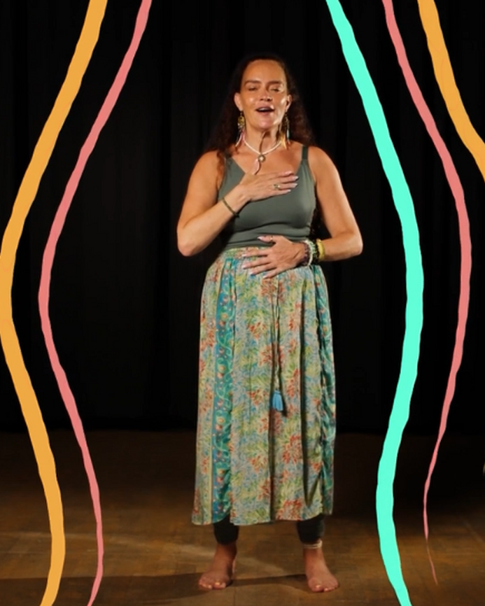 Michelle Hamilton standing in a studio, with illustrations of colourful lines around her, she has one hand on her heart and one hand on her stomach