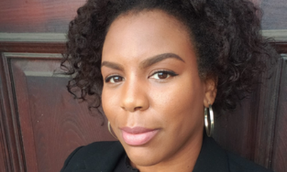 a headshot of Nola Sterling, standing in front of a door wearing a black blazer