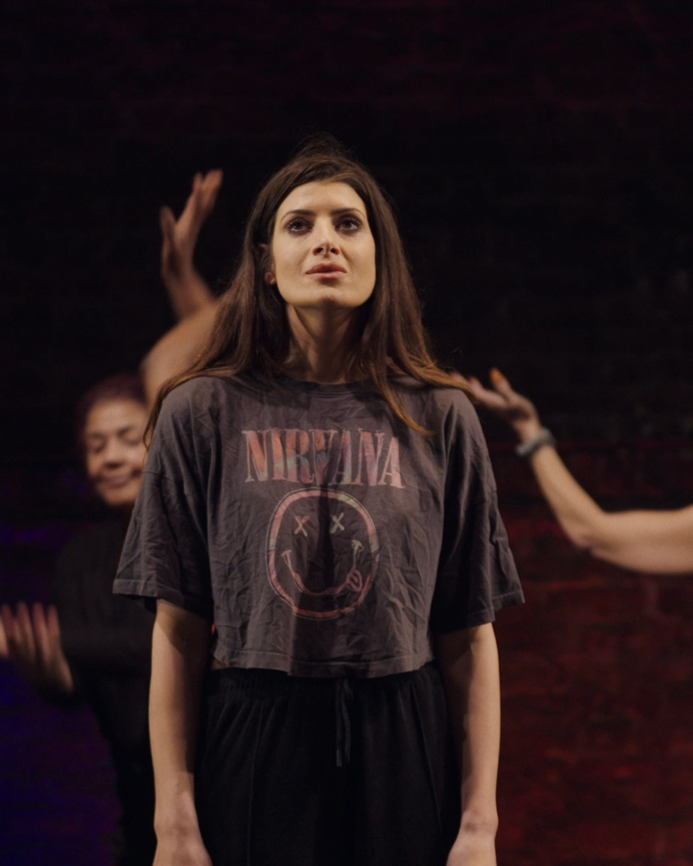 Natasha on a stage, looking upwards, surrounded by Clean Break Members