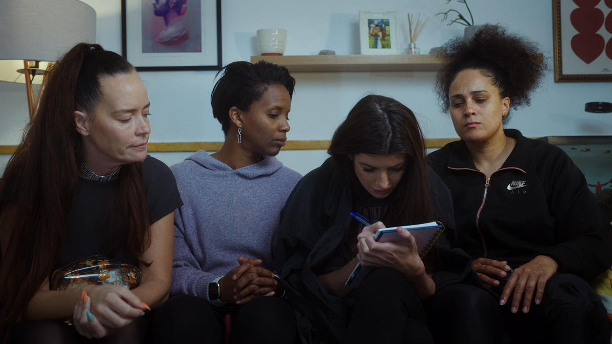 The cast of Hope sitting on a sofa, Natasha is writing in a notebook, the rest are looking over to see what she is writing