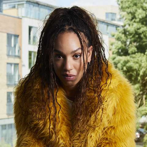 Pearl Mackie as Baby, wearing a yellow fur coat with a defiant yet vulnerable look on her face, sitting in front of a window