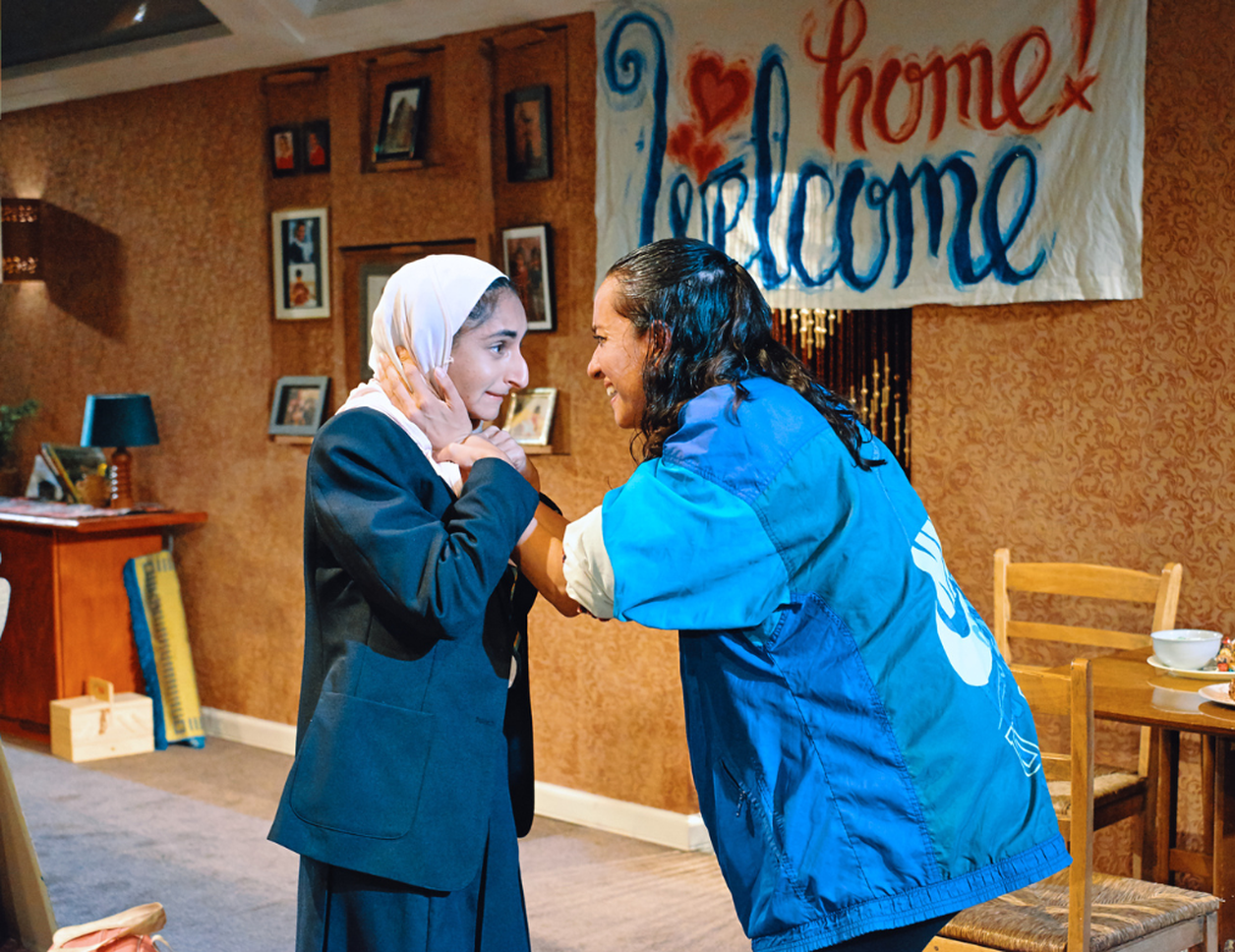 a production photo for Favour, Aleena is holding Leila's face in her hands and looking at her. In the background a banner reads 'Welcome home