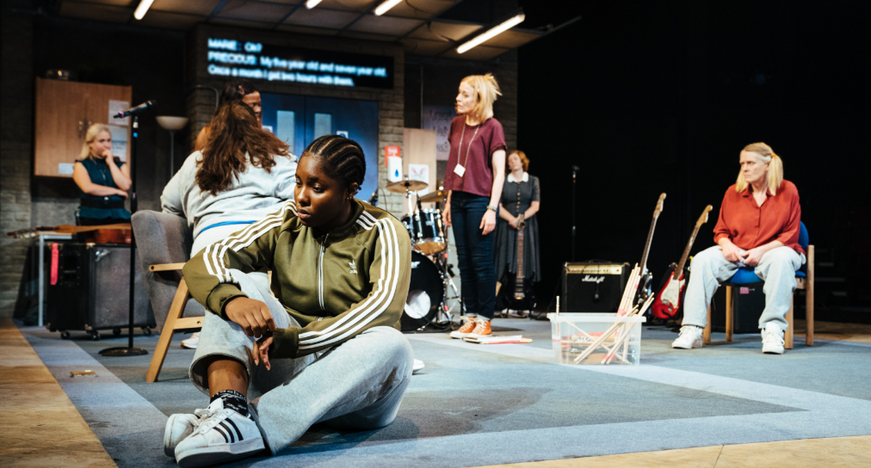 The Typical Girls cast on stage, Lara Grace Ilori is closest to the camera, she is sat on the floor with her legs crossed, looking at the floor.