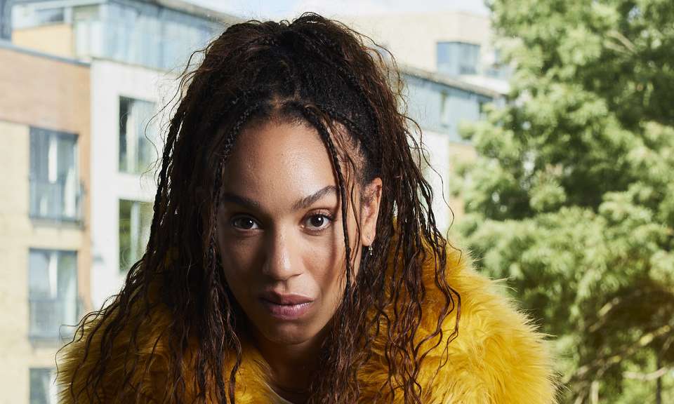 Pearl Mackie as Baby, wearing a yellow fur coat with a defiant yet vulnerable look on her face, sitting in front of a window