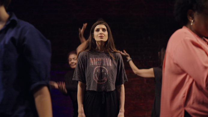 Natasha on a stage, looking upwards, surrounded by Clean Break Members