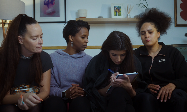 The cast of Hope sitting on a sofa, Natasha is writing in a notebook, the rest are looking over to see what she is writing