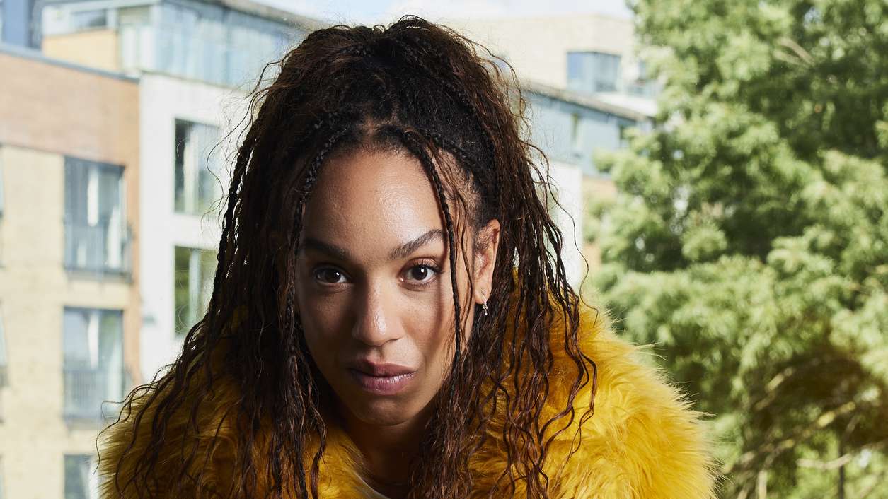 Pearl Mackie as Baby, wearing a yellow fur coat with a defiant yet vulnerable look on her face, sitting in front of a window