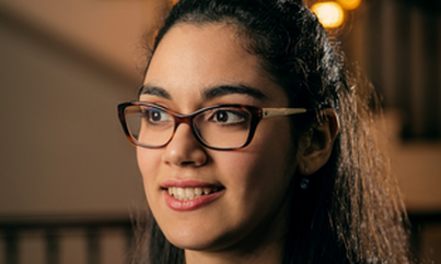 A headshot of Sophie Dillon Moniram, it is close up to her face, she is smiling and wearing glasses, she has long brown hair, the lighting is warm.