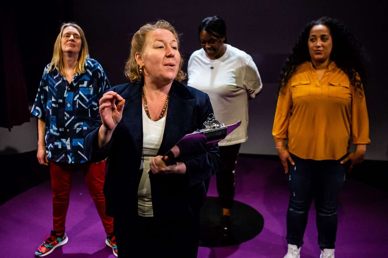 a production photo from Inside Bitch of four women on stage, looking forwards, one of them is holding a clipboard