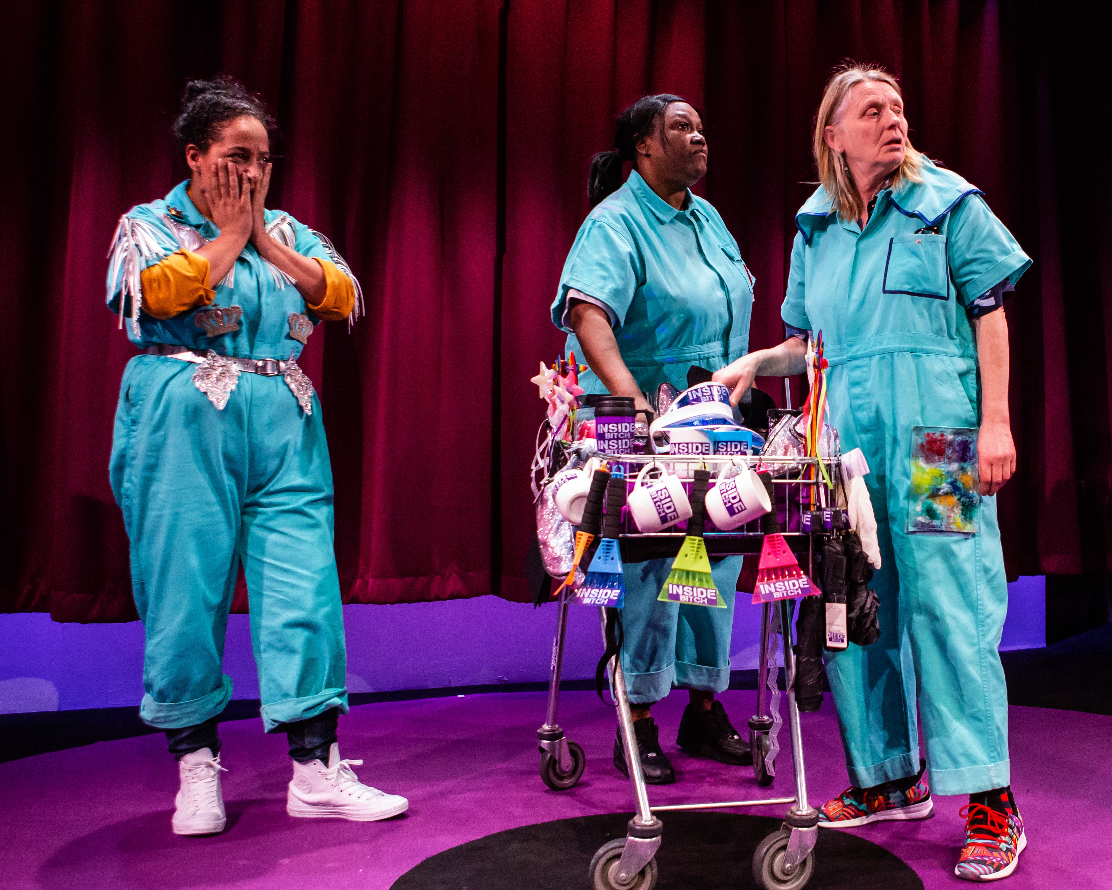 An image from Inside Bitch, three characters are on stage with a trolley full of 'Inside Bitch' merchandise,
