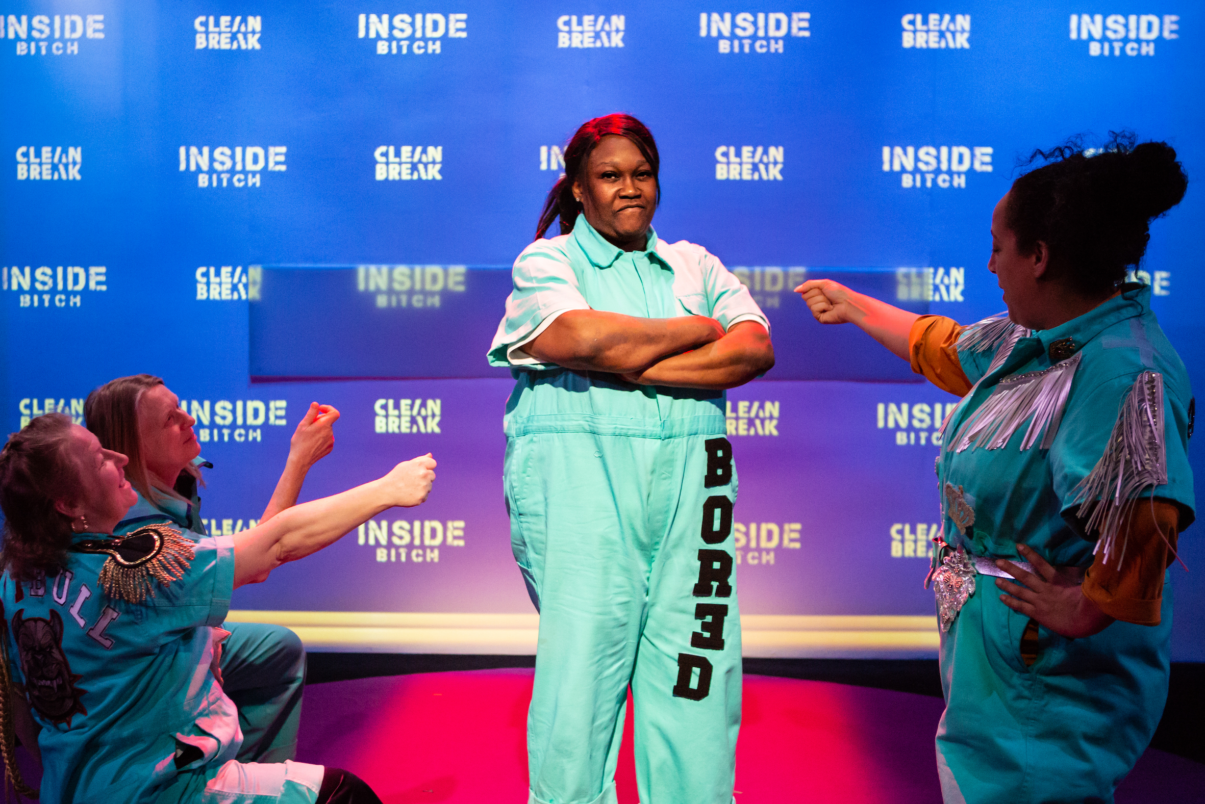 A photo of Jennifer Joseph in Inside Bitch. She is standing up wearing a turquoise jumpsuit which says 'bored' on the leg.