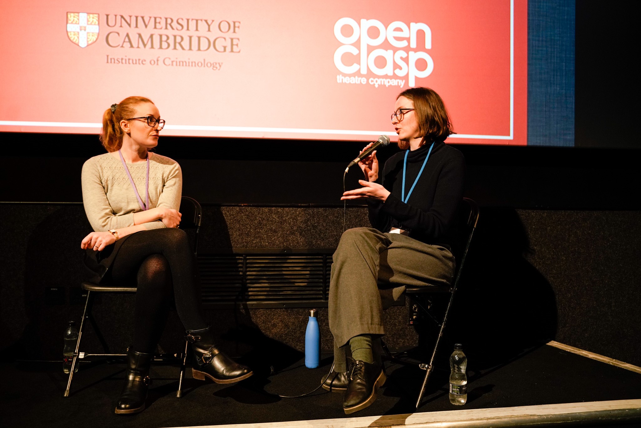 Clean Break Producer Maya Ellis is shown speaking into a microphone on stage with Open Clasp Senior Producer Carly McConnelly